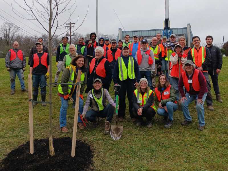 A large group of volunteers pose near a newly-planted tree on a gently-rising slope at a WPC tree planting event.